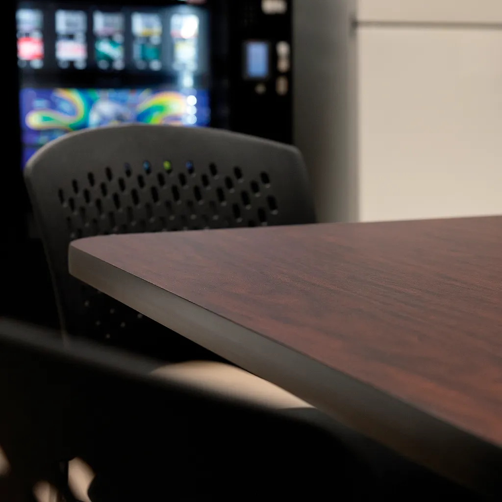 Close-up of table's dark brown wood grain top and black edge, with chair and vending machine in background
